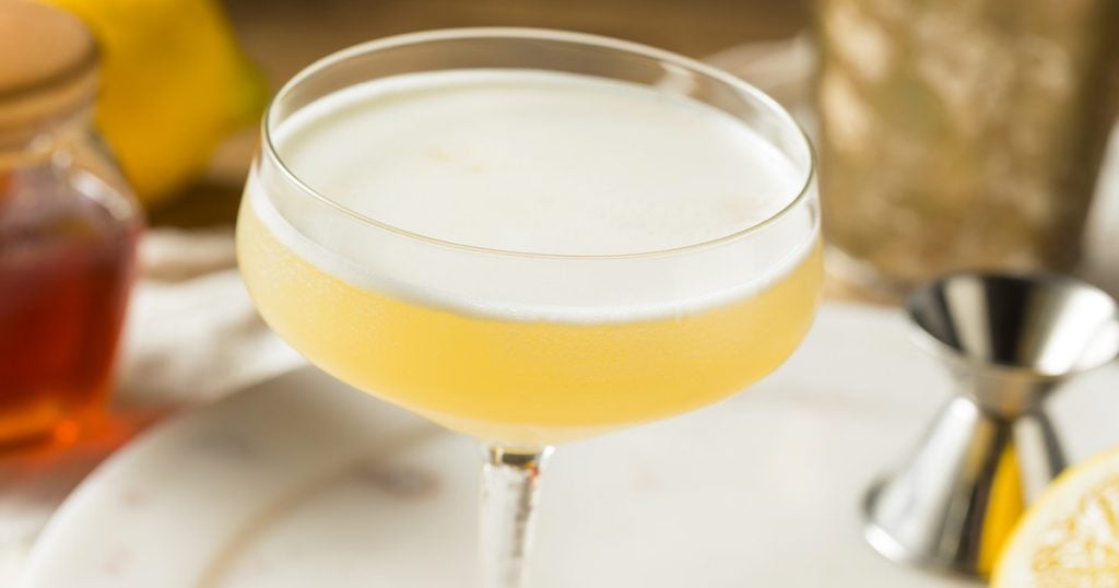 A Bee's Knees Cocktail in a couple glass with honey, lemon, and a jigger in the backdrop