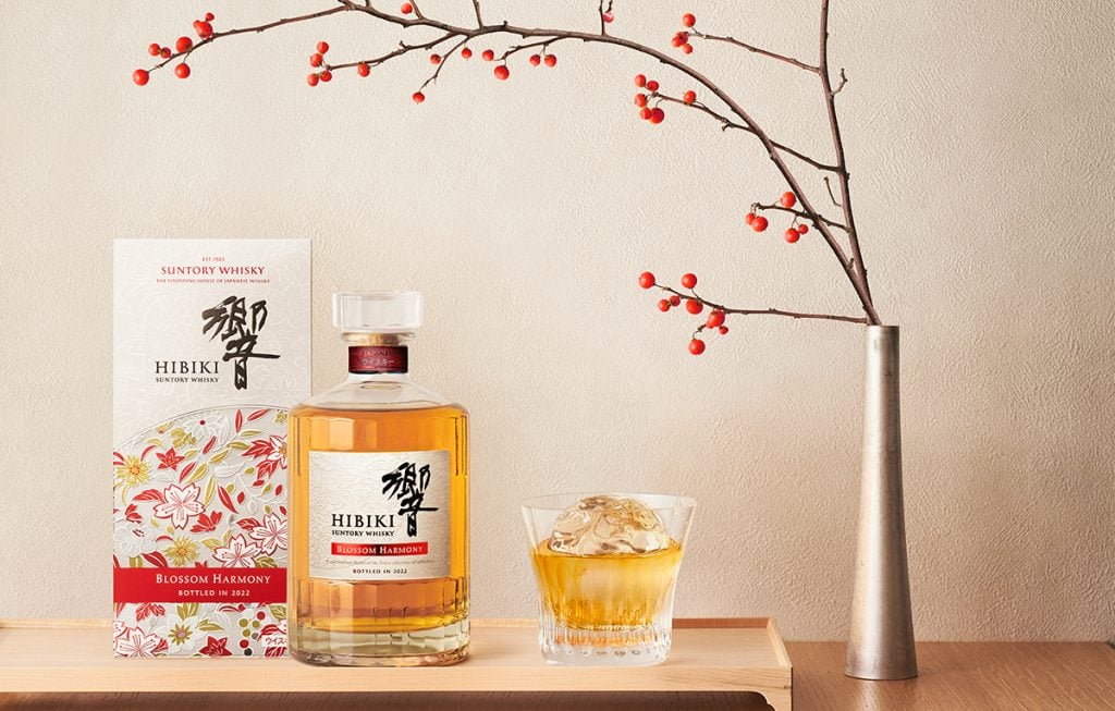 A bottle of Hibiki Blossom Harmony 2022 with the presentation box, a glass of whisky with ice, and a plant