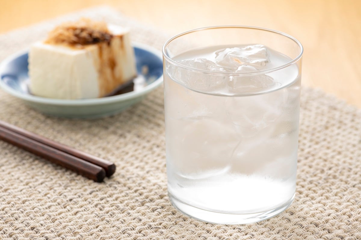 A close up of a glass of awamori over ice, next to some chopsticks, with a plate of cold tofu in the background
