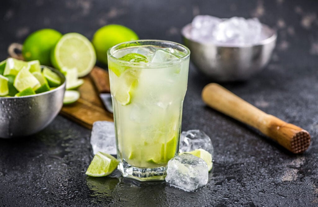A glass of Caipirinha with cocktail ingredients and limes around it