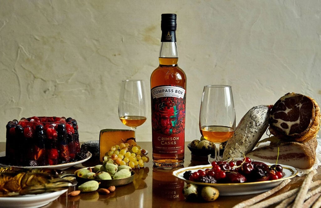 A bottle of Compass Box Crimson Cask with glasses of whisky and a spread of food and drink such as dried fruit and cake