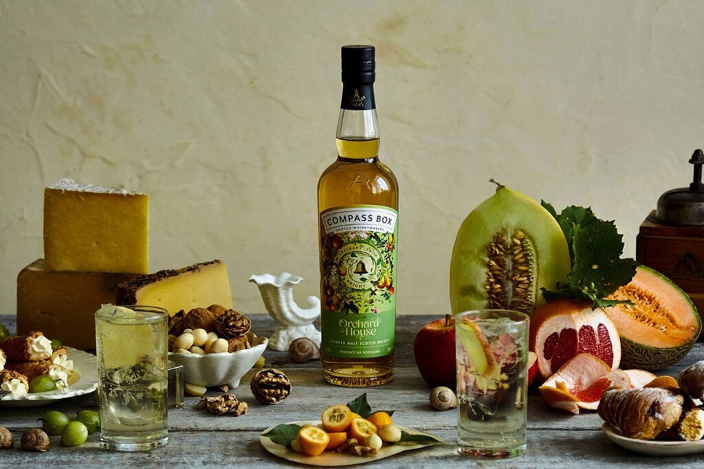 A bottle of Compass Box Orchard House with a spread including cheese, fruit, and glasses of whisky