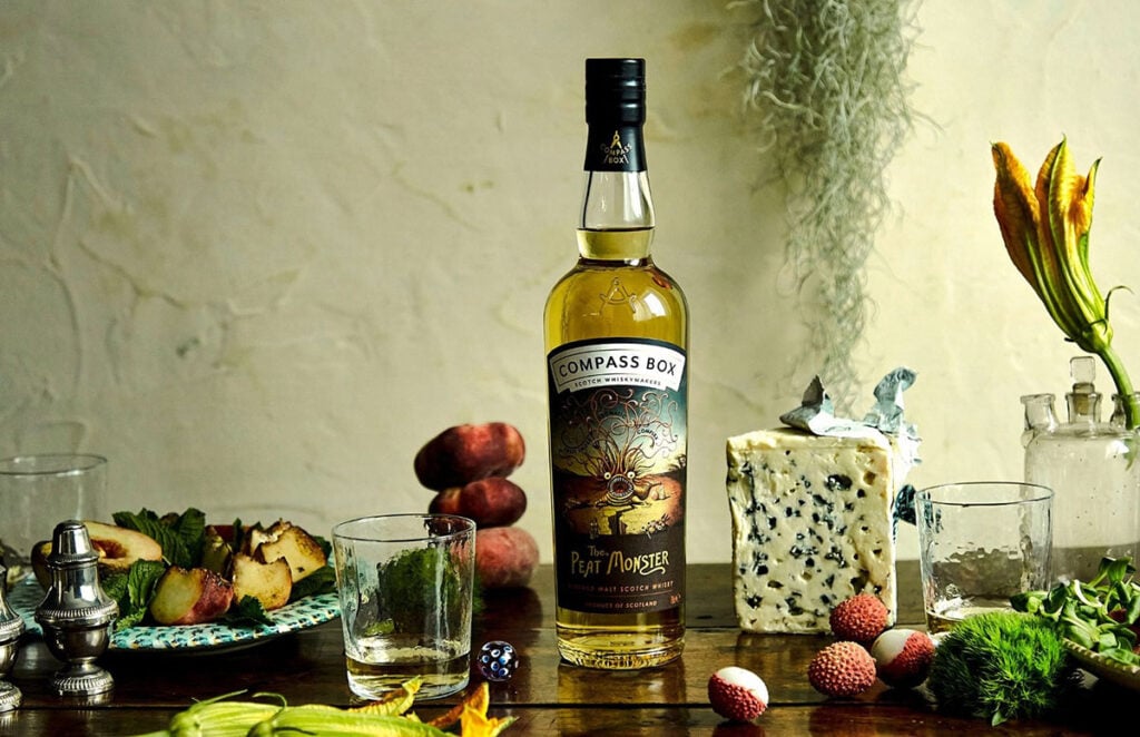 A bottle of Compass Box Peat Monster with glasses of whisky and a spread of food and drink such as blue cheese and fruit