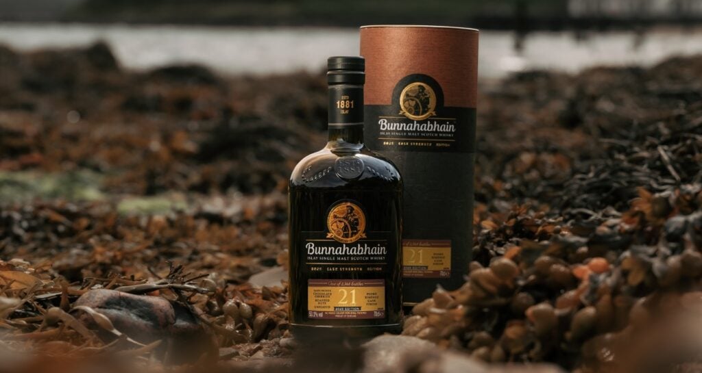 A bottle of Bunnahabhain 21 Year Old Cask Strength - 2025 on the beach, surrounded by seaweed.