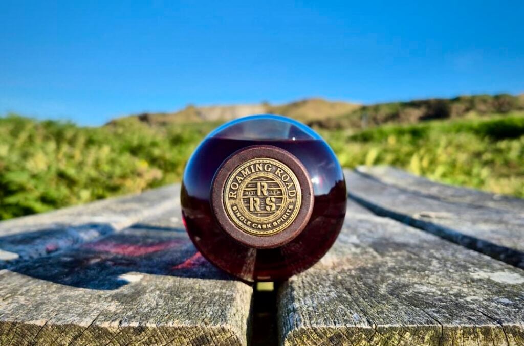 A bottle of Roaming Road whisky on a bench in a field