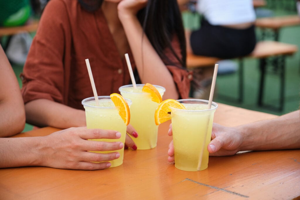 Hands holding glasses of fresh lemonade with orange slices and paper straws at a summer party