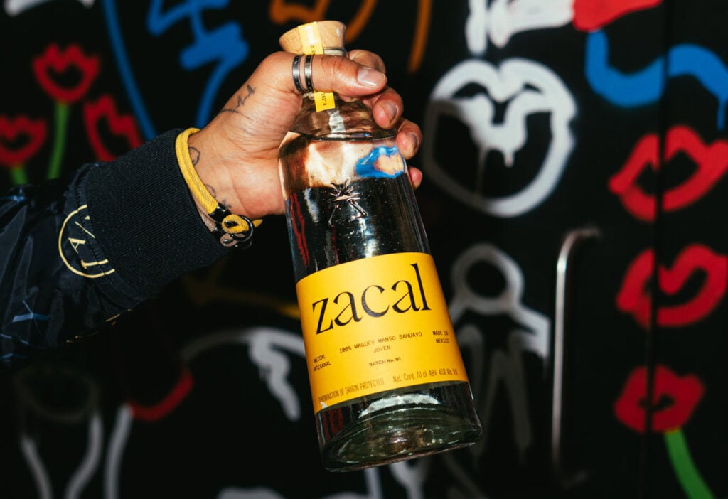 A bottle of Zacal mezcal in a person's hand in front of a graffitied wall