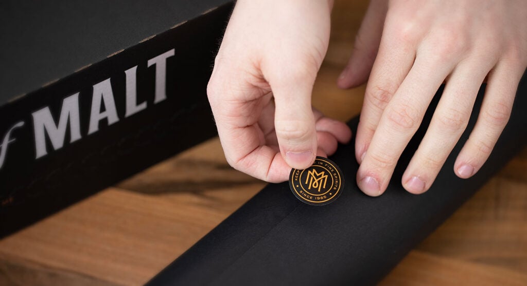 A Master of Malt Mystery Drop Box next to somebody putting a label on a bottle