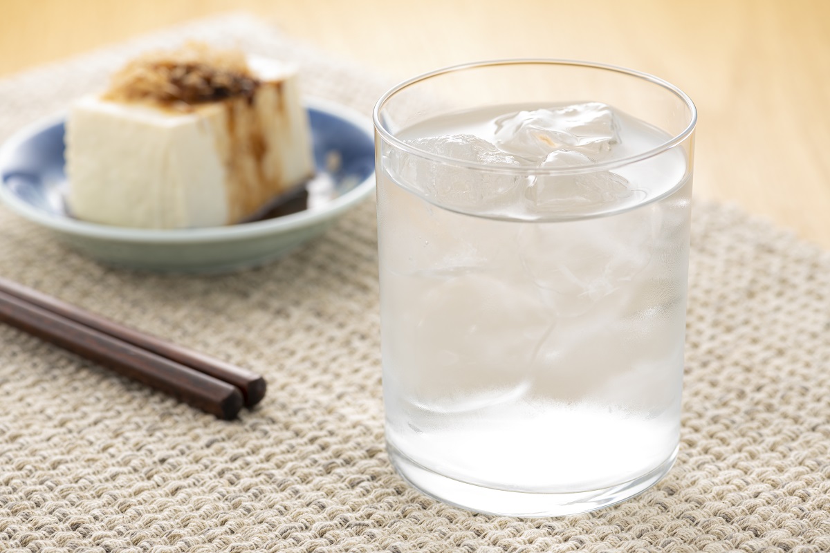 A close up of a glass of awamori over ice, next to some chopsticks, with a plate of cold tofu in the background