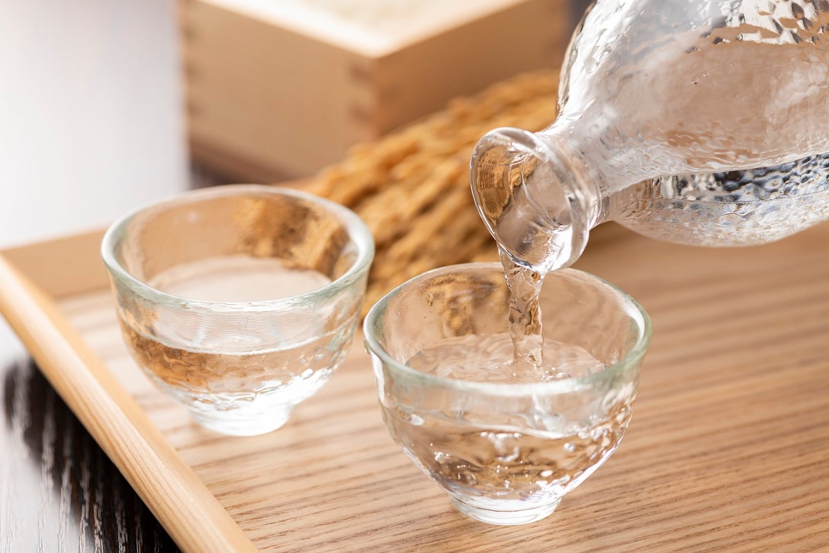 Two small glasses full of clear liquid. Sake is being poured from a clear glass bottle into one of the glasses