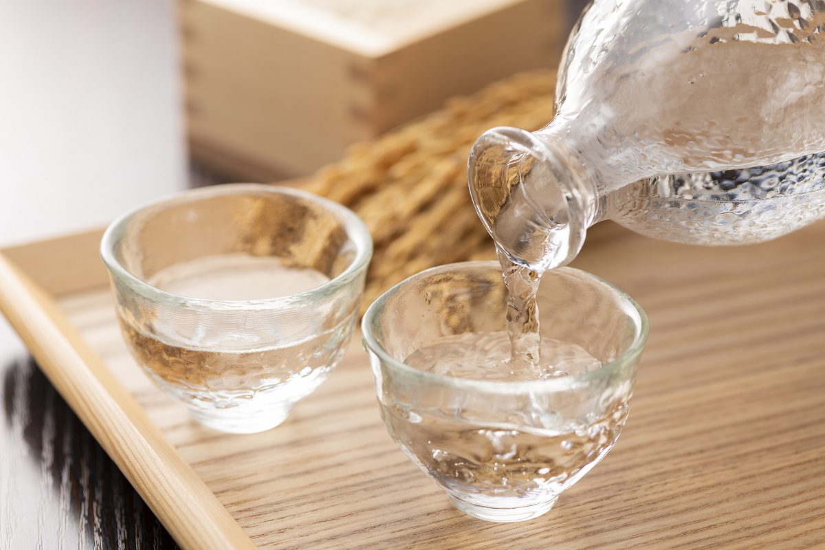 Two small glasses full of clear liquid. Sake is being poured from a clear glass bottle into one of the glasses
