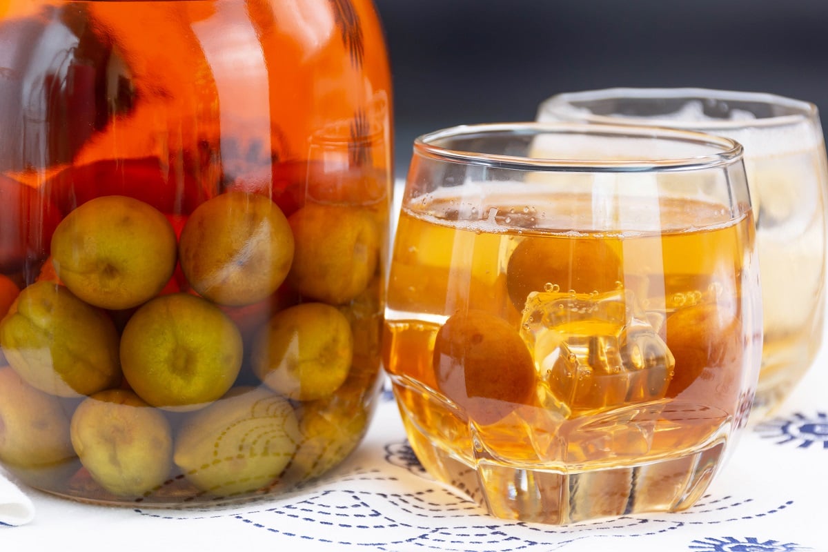 A table with a jar of ume, submerged in amber liquid. Next to it is a glass of umeshu on the rocks, with a few plums in there too.