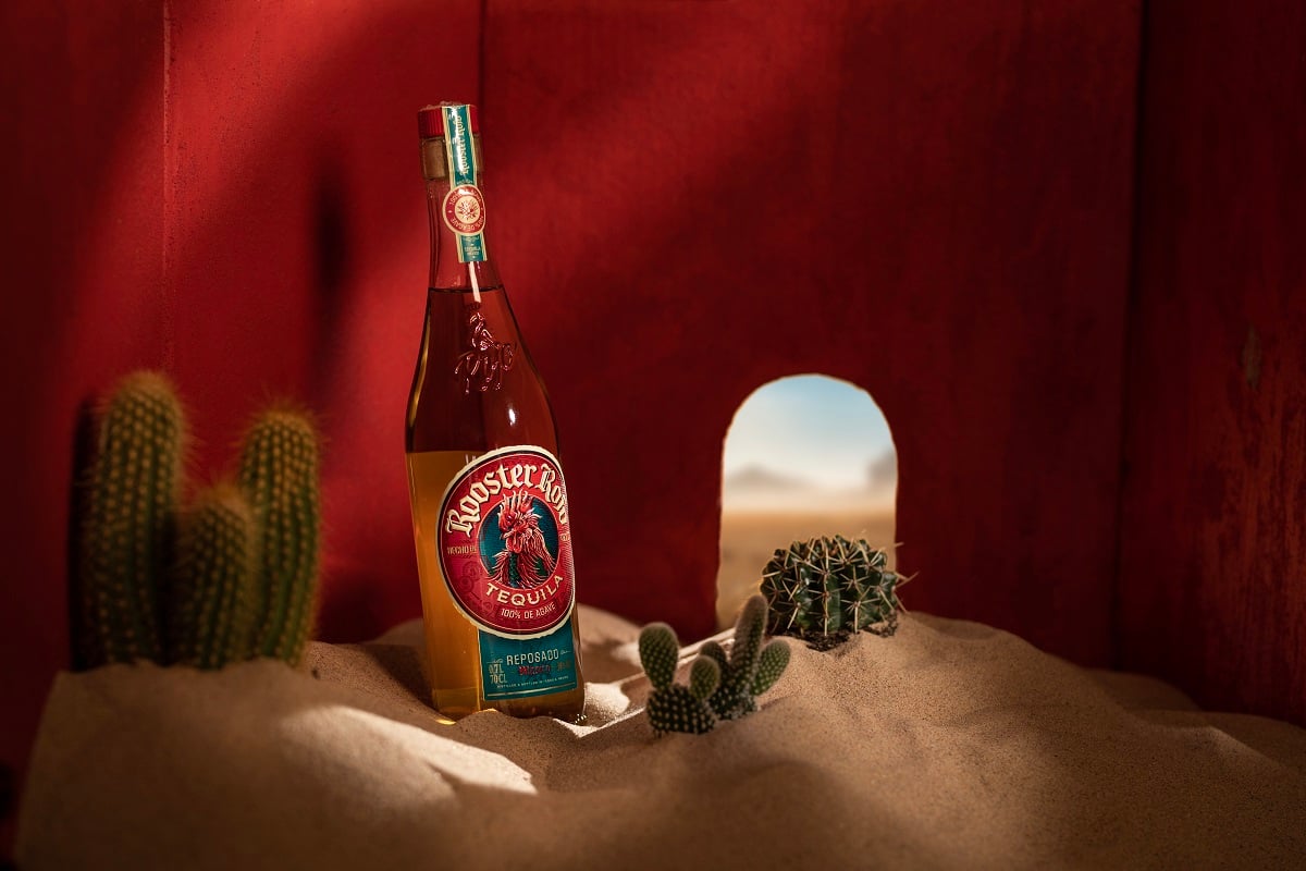 A bottle of Rooster Rojo Reposado Tequila in a room with red walls that's filled with sand and cacti