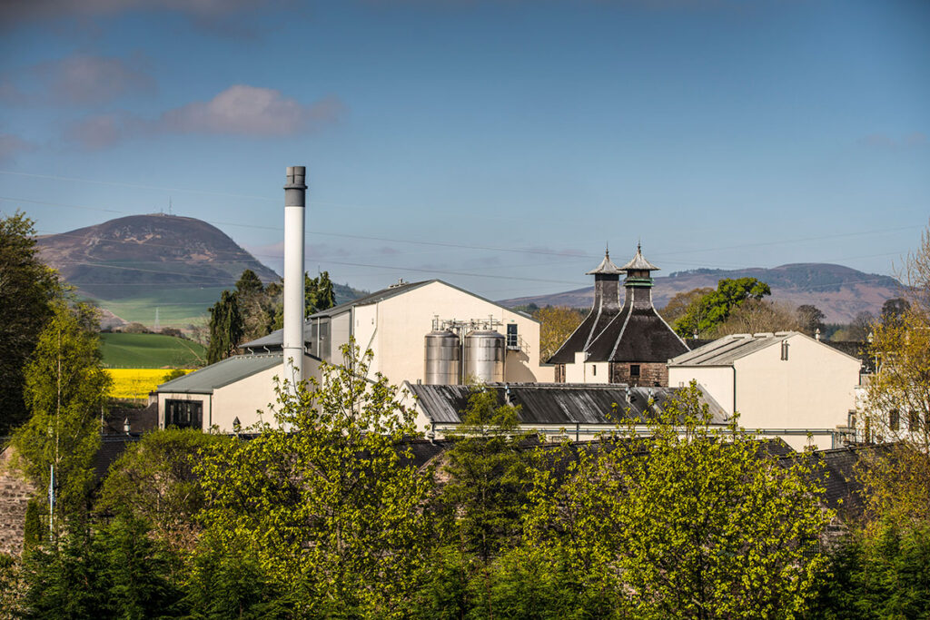 The Glen Ord Distillery pictured in its Highland surroundings with mountains, trees, and fields.
