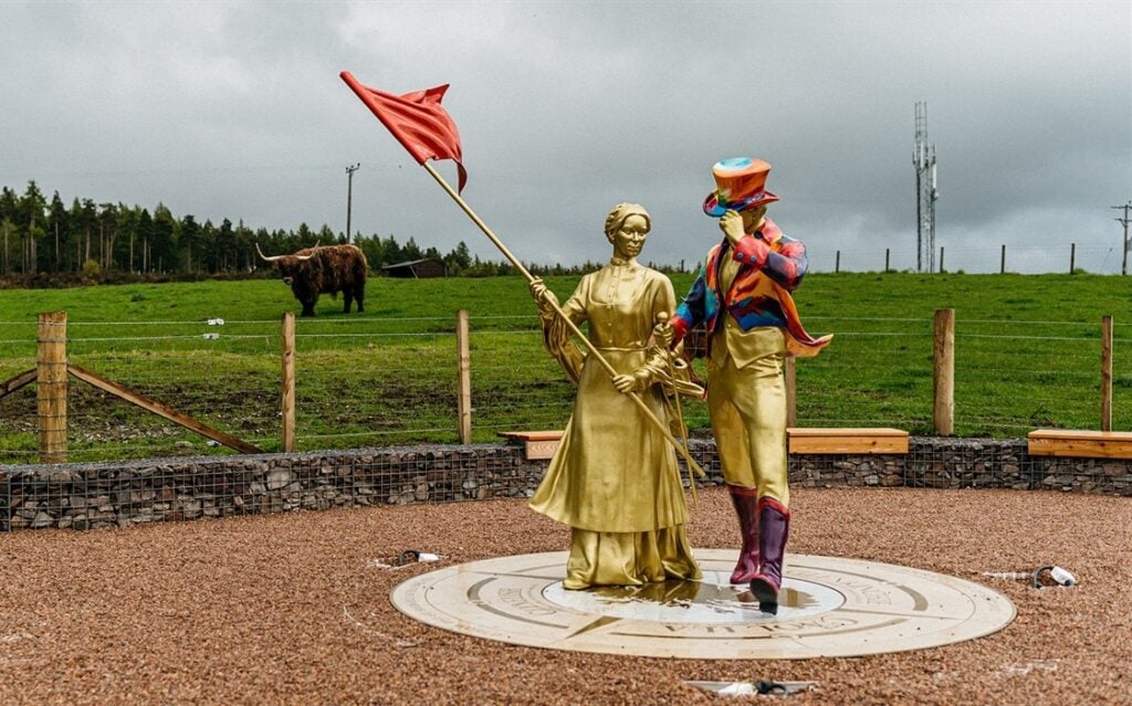 Helen Cumming – raising her famous red flag – alongside the iconic Johnnie Walker Striding Man. The statues sit just outside Cardhu, the Speyside Home of Johnnie Walker